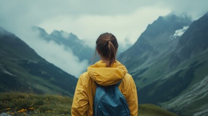 Fototapeta premium Woman wearing a yellow raincoat and blue backpack hiking in the Tyrol mountains, gazing at stunning peaks and clouds, vibrant green landscape.
