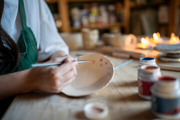 Unrecognizable ceramist applies flower paint to ceramic plate, making artistic author pattern. Creative occupation, handmade dishes, artisan clay production, unique craftsmanship, painted tableware