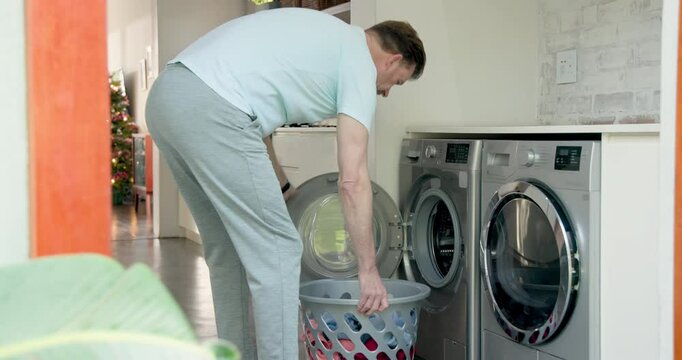 Senior man doing laundry at home, loading clothes into washing machine - Powered by Adobe