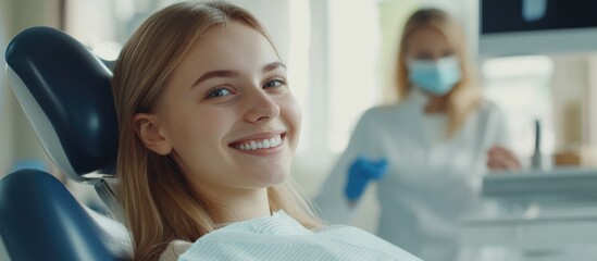 Caucasian young woman smiling joyfully in dental chair post-treatment with dental assistant in background wearing protective gear in modern clinic setting
