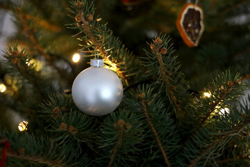 Elegant, pearl white christmas bauble hanging on the fir branch lit by the light chain. Blurry dried orange slice decoration in the background