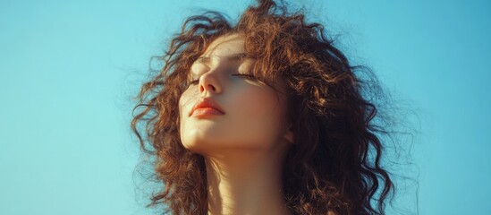Obraz premium Beautiful young woman with curly hair against a bright blue sky, serene expression, large empty space for text, fashion photography concept.