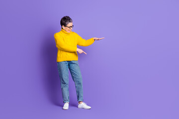 Full length photo of charming positive woman wear yellow sweater showing arm pointing empty space isolated violet color background