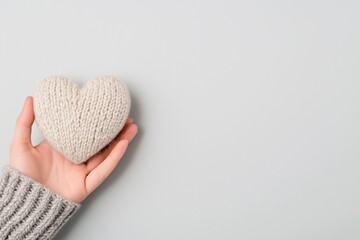 Fototapeta premium A soft, human hand holding a heart-shaped object knitted from cream-colored yarn. The background is a clean, neutral tone, enhancing the delicate and cozy aesthetic