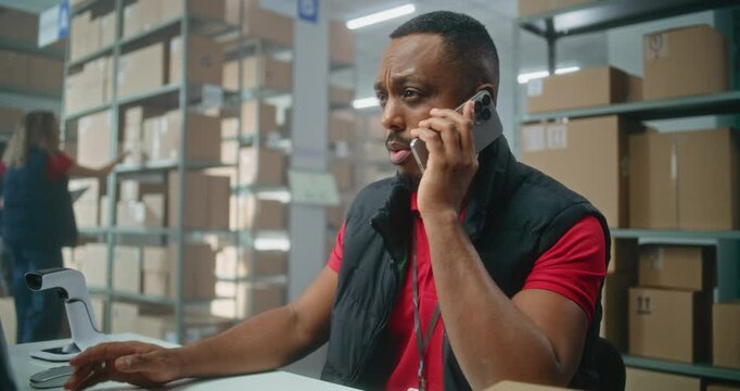 Warehouse retail facility of E-commerce store or postal service: African American logistics coordinator talks by phone, uses computer. Sorting center workers checking cardboard boxes for delivery.