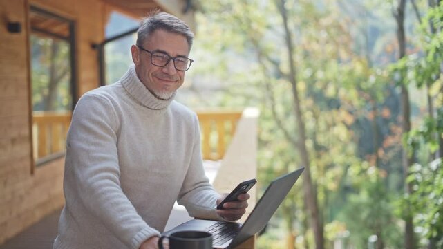 Mature businessman using laptop and smartphone on wooden terrace of mountain chalet, remote working from peaceful nature. Portrait of happy mid adult man wit grey hair and glasses, smiling.
