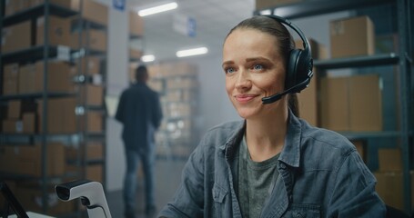 Warehouse of E-commerce online store or post office: Female customer support specialist in logistics talks by headset with client, uses computer. Sorting center employee checking boxes for delivery.