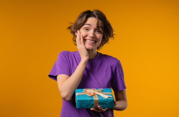 Fototapeta premium Grateful woman with gift box on yellow. Confused lady smiling, Present, surprise