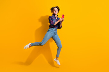 Full length photo of clever optimistic woman dressed plaid jeans shirt jeans fly holding smartphone isolated on yellow color background