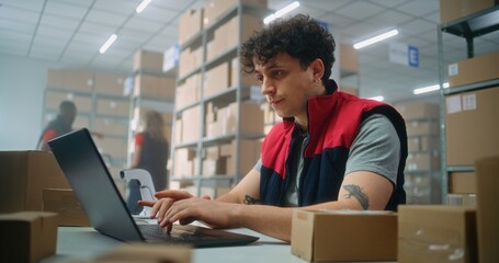 Parcel processing specialist scans package using barcode scanner, prepares for shipping to customer, works on laptop. Warehouse employees carrying boxes. E-commerce store or postal service storage.