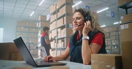 Portrait of female logistics coordinator wearing headset talking to customer, working on laptop in postal service or online store storage, looking at camera. Sorting center employees checking boxes.