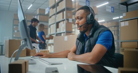 African American logistics coordinator wearing headset chats with customer on call, uses computer,...