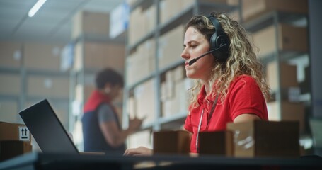 Female logistics coordinator wearing headset chats with customer on call, uses laptop, works in storage of post office or E-commerce store. Sorting center workers checking cardboard boxes. Slow motion