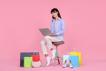 Internet shopping. Happy woman with laptop sitting on stool among colorful bags against pink...