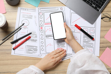 Website designer holding smartphone at wooden table with wireframes and stationery, top view
