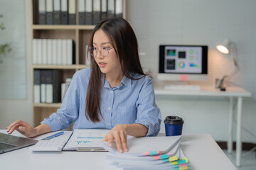 Young Asian businesswoman analyzing financial paperwork and working with laptop computer at her...