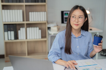 Young Asian businesswoman holding a cup of coffee and working with laptop computer and analyzing financial chart document at office desk, thinking about new ideas for her business