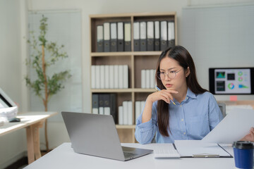 Young Asian businesswoman holding a pen and focused on her laptop, analyzing financial documents and strategizing for her project in a modern office environment