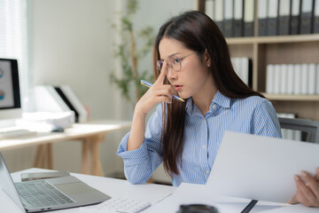 Young asian businesswoman adjusting eyeglasses and thinking about accounting using laptop and documents, working hard at office desk with laptop computer, financial accounting concept