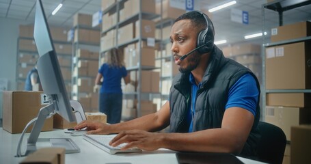 African American logistics coordinator wearing headset chats with customer on call, uses computer,...