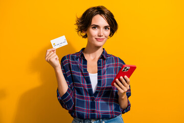 Photo of intelligent cheerful woman dressed checkered shirt hold smartphone credit card isolated on vibrant yellow color background