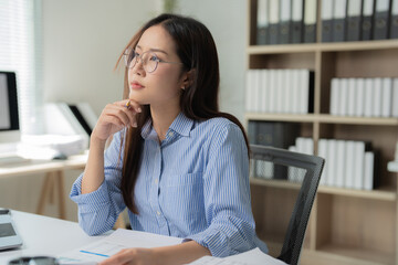 Young Asian businesswoman wearing glasses, deep in thought while holding a pen, is diligently...