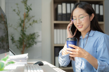 Asian businesswoman making a phone call while sipping takeaway coffee, seated at her desk in a...