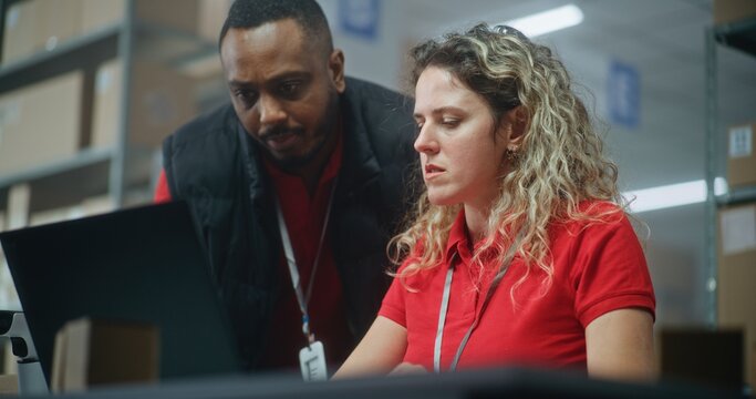 African American warehouse associate with tablet computer comes to female logistics coordinator working on laptop, consults about delivery online orders to customers. Distribution or sorting center.