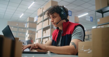 Customer support specialist in logistics wearing headset talks to customer on call, uses laptop,...