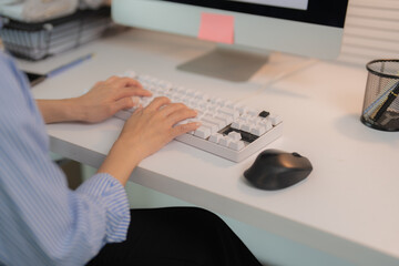 Close-up of female hands typing on a white keyboard on a white desk in a bright office, with a computer monitor in the background and a mouse nearby, suggesting a productive workday