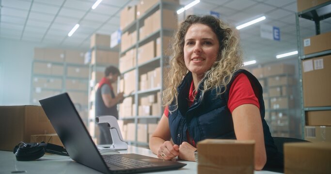 Portrait of female post office employee scanning cardboard parcel with barcode scanner, using laptop, looking at camera. Sorting center workers checking boxes for delivery. Warehouse of online store.