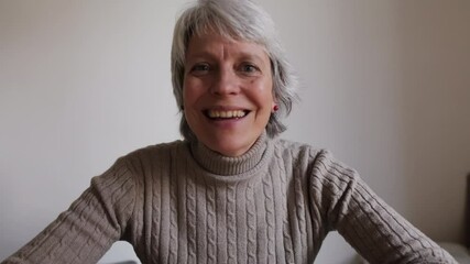 An older woman with grey hair smiles and talks during a video conference from home. She is happy and engaged in conversation.