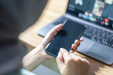 A Young business woman interacts with a smartphone while sitting in front of a laptop, suggesting multitasking in a digital environment.