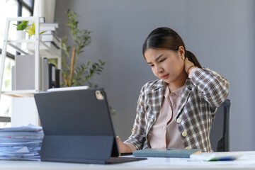 Asian businesswoman suffering from neck pain while working on a tablet in the office, concept of occupational health problems