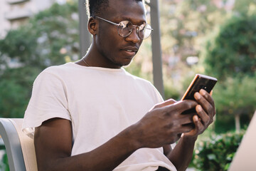 Fototapeta premium Concentrated african american hipster guy in eyewear for vision correction reading notification on smartphone, serious dark skinned businessman checking mails on mobile phone on remote job