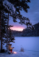 Man sitting and drinking coffee near a campfire in a winter landscape with the glowing sky after the sunset behind