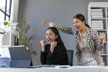 Two euphoric businesswomen screaming with joy while looking at a tablet in the office, celebrating...