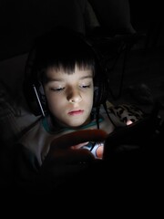 Child Using Phone in Bed. A young boy with short dark hair is lying in bed, wearing headphones and looking intently at a smartphone. The room is dark, with only the light from the phone screen 