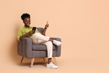 Cheerful woman with smartphone sitting in armchair and pointing at something on beige background....