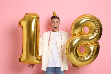 Coming of age party - 18th birthday. Emotional young man with number shaped balloons and hat on pink background