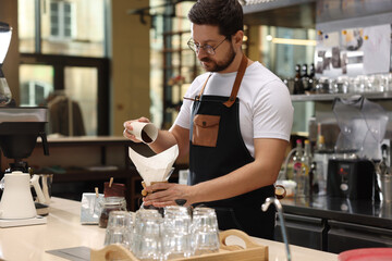 Barista pouring ground coffee into glass coffeemaker with paper filter at table in cafe