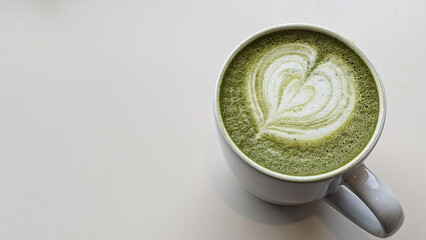 A matcha latte with heart-shaped foam art in a white ceramic mug, placed on a table with greenery. Concept of matcha, heart, latte, cup, foam, drink, table, green, white, pattern.