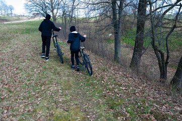 Naklejka premium Two individuals, dressed in dark clothing, walk alongside their bicycles on a leaf-strewn path. Trees line the trail in a park-like setting.