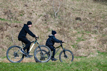 Fototapeta premium Two young people on mountain bikes cycle along a grassy path. The background features a gently sloping hillside. The scene suggests a sunny day and an outdoor recreational activity.