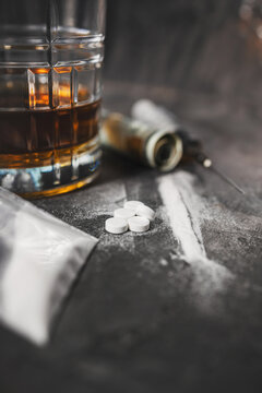 White narcotic pills, an alcoholic drink in a glass, a syringe with a dose of heroin, a line of cocaine powder and US dollar bills on a dark background. Concept of addiction and bad habits