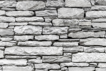 Close-up view of an aged stone wall, with irregularly shaped stones stacked horizontally to create a rugged facade or sturdy structure. The stones are light gray in color.