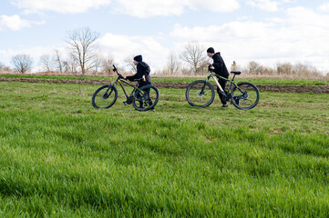 Obraz premium Two individuals in dark clothing stroll across a verdant field, pushing their mountain bikes. The scene is peaceful and suggests a relaxing outdoor experience.