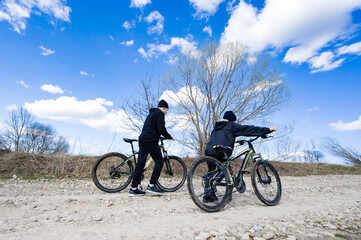 Obraz premium Two young people take a break from cycling on a dirt road. The bright blue sky and sparse trees frame the scene, suggesting a spring or autumn day.