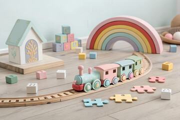 A pastel-colored toy train set on a wooden floor, surrounded by puzzle pieces, building blocks, and a rainbow-shaped decor piece. The minimalist yet playful composition highlights creativity and fun