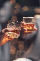 Cheers to Connection: Two hands clink glasses of amber liquor, the warm glow of the beverage reflecting the blurred lights of the background in a toast to friendship, celebration.